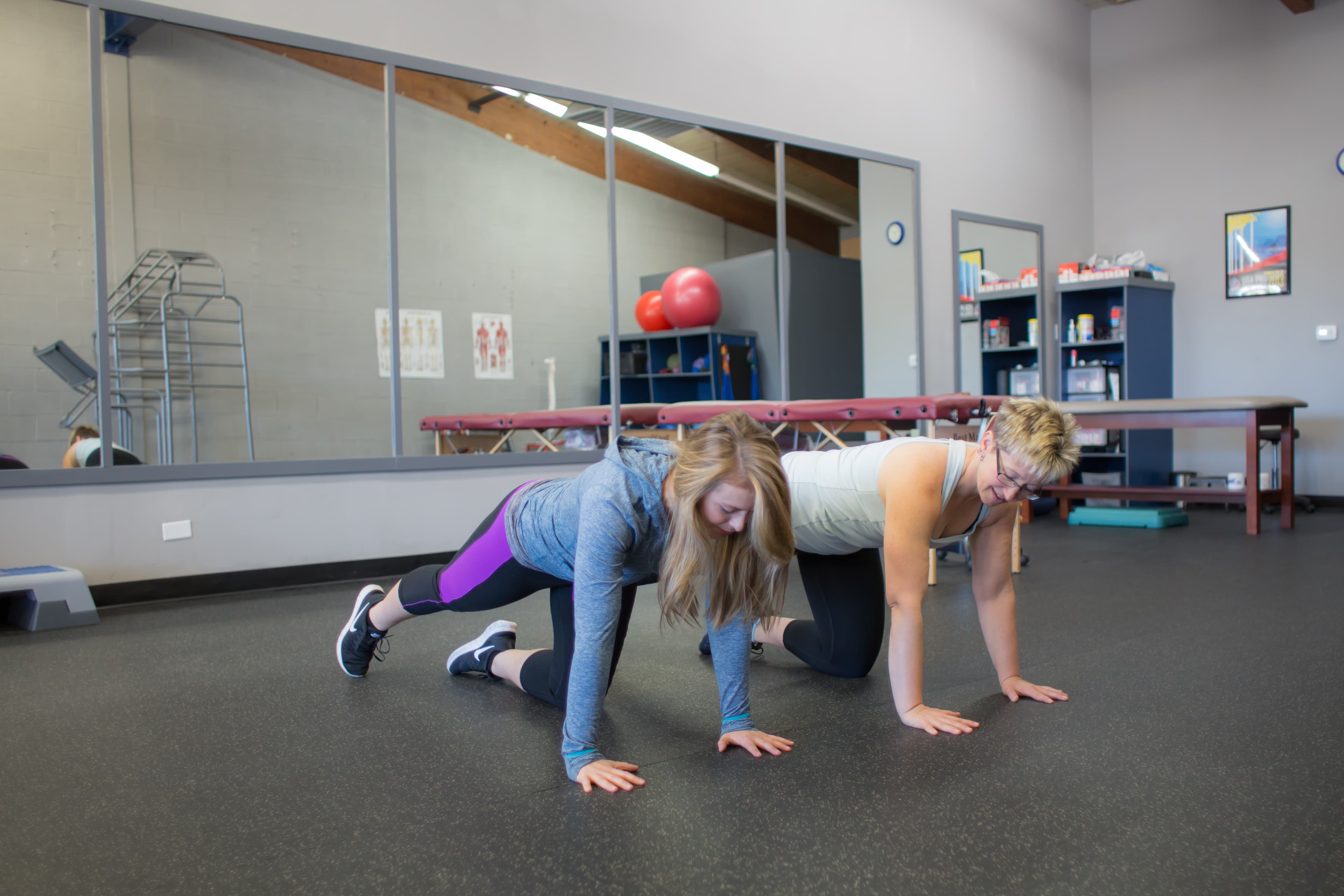 Two women on their hands and knees doing a guided exercise in the clinic gym, beside a mirror wall
